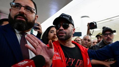 Tarrio in sunglasses and a red sweater gestures towards a microphone in the middle of a crowd of people.