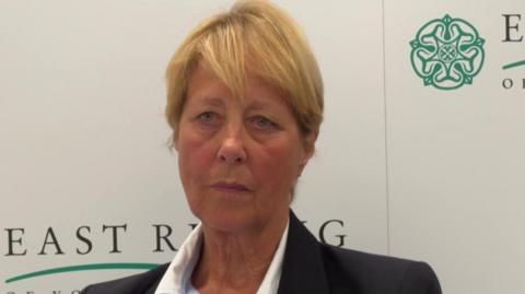 Conservative councillor Victoria Aitken. Woman with short blonde hair wearing a dark blazer and a striped shirt. She is sat in front of a board with East Riding of Yorkshire Council across it.