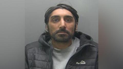 Custody photo of Iltaf Hussain, 40. He is looking straight ahead at the camera with a grey background. He can be described as having shoulder length hair, with a moustache and short beard. 