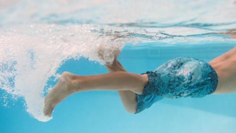 A young boy swimming under water in a pool, he has blue swim shorts on and is kicking in the water.