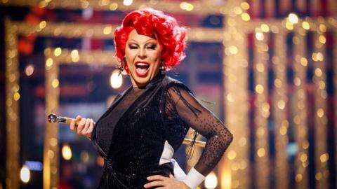 La Voix wears a short red wig in curls. They are wearing a black glittery and sheer dress and holding a cane with a glittery blurred background behind them