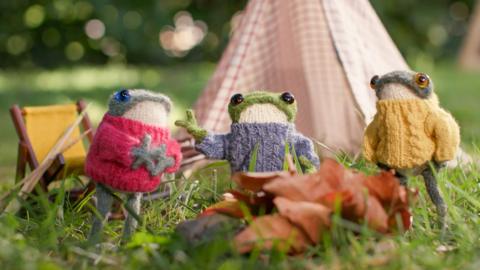 Three froglets wearing jumpers, sat around a camp fire.