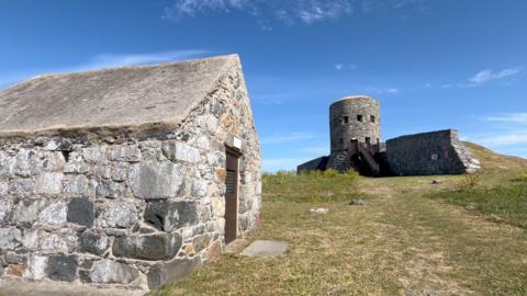 A small stone hut with a slanted concrete roof sits on a grassy hill, with a larger round stone tower behind it. The tower has narrow windows, an attached wall section, and a wooden staircase leading to its entrance. Both structures stand under a clear blue sky. 