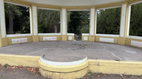 An empty bandstand that has been painted yellow and white. There is a large flat area for bands to before on and trees are seen to be surrounding the structure. There are light pinkish marks on the base of the bandstand where the St George's cross had been painted. 