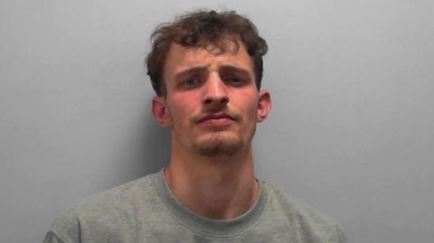 Frankie Lee Morrison's police mugshot - against a plain background, a young man stands wearing a grey jumper. He has short curly brown hair and a goatee beard.