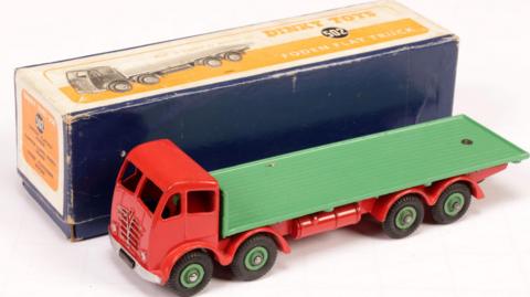 A long, green and red toy truck is placed beside its cardboard box. The cab is red and has holes for windows and grey detailing depicting the grill and headlights. The wagon in the back is a single piece of green metal. The box is yellow and blue. text on it reads: "Dinky Toys 502 Foden Flat Truck."