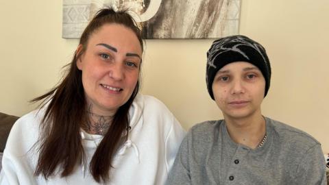 A woman with scraped-back long dark hair is wearing a white hoodie and sitting on the sofa. She has a nose ring and a tattoo on the front of her neck. On her left there is a teenage boy wearing a grey Henley shirt and a black beanie hat.