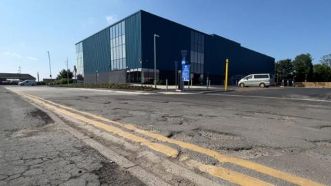 A brand new leisure centre building with the road in front of it showing a number of potholes and damage to the pavement. 