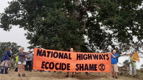 A banner reading National highways ecocide shame at the site of the tree with protesers