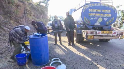 People wearing caps and mufflers fill water in their buckets and drums from a public water tanker in the Rajouri district.