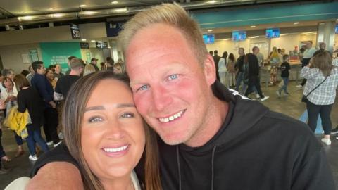 A couple take a selfie photo - on the right is a man with short blonde hair and blue eyes wearing a black hooded sweatshirt. He has this arm around a shorter woman with long brown hair wearing a white top. They are both smiling at the camera.