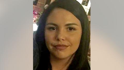 Alice Webb is smiling at the camera, she has dark hair and a piercing in her lower lip. She stands in front of an illuminated bar.
