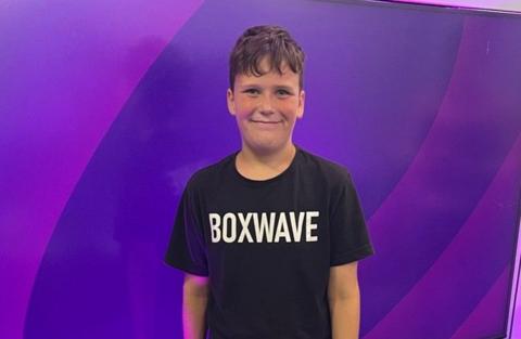 A ten-year-old boy wearing a black t-shirt that reads "Boxwave" standing in front of a purple screen. He is smiling and has short brown hair.