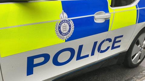 The side of a Guernsey police car with the Guernsey Police crest in the centre of the car door. The car is coloured white with neon yellow and blue coloured livery. 
