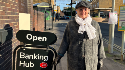 Woman with hat and scarf by a sign which says 'open' and 'banking hub'