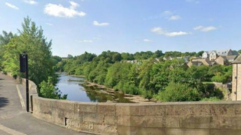 A view over County Bridge in Barnard Castle over the River Tees. Both sides of the river are covered in greenery and trees. A traffic light can be seen on the bridge to the left.
