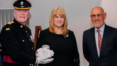 A man in military dress and white gloves shows a glass plague towards a camera. He is stood next to a woman with light hair in the centre and a man with a pinstriped suit on the right.