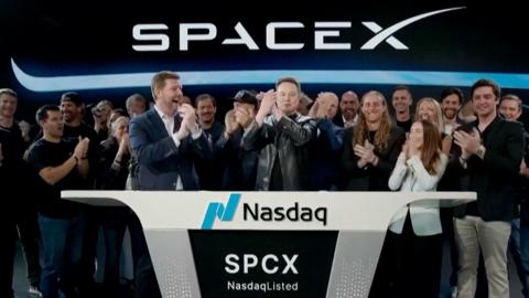 Elon Musk wearing a black leather jacket, stands in the center of crowds and in front of a SpaceX sign.