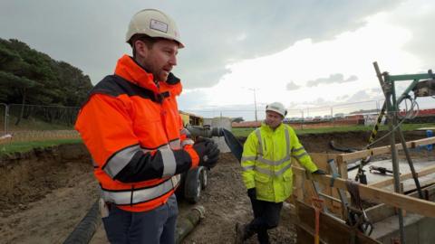 Tom Le Gallais at the site wearing a orange hi-vis jacket and a cream hard hit. Another man is also at the site wearing a yellow hi-vis jacket and safety helmet. The site is muddy.