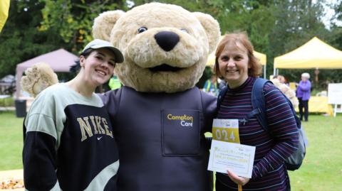 Two women stand either side of someone dressed as a large teddy bear. The teddy bear costume says "Compton Care" on it in yellow and white letters