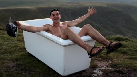 A man is bathing in a bathtub posing for a camera while on a mountain summit. He has short dark hair, holds a black cap in one hand, and is wearing flip flops. 