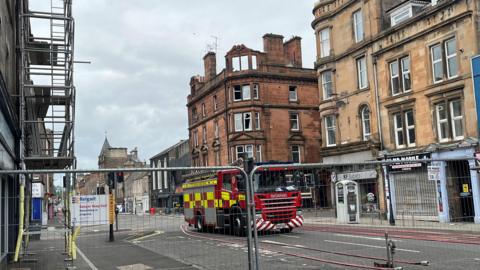 A burned-out building is cordoned off by a fence, with a fire engine in the centre of the picture 