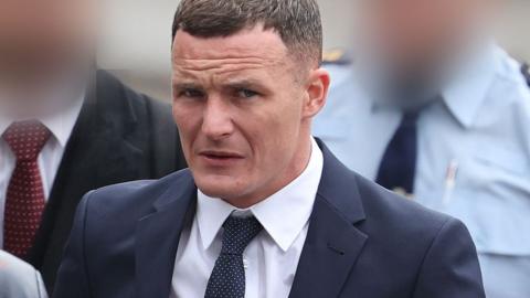 Kielan Mooney pictured at a previous court hearing. He has short brown hair and is wearing a suit and tie. Two people, who have their faces blurred, can be seen behind him. 