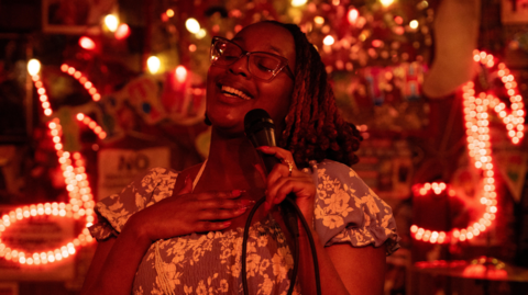 Edna Nicole Luckett sings at a microphone, her hand on her heart, with light-up signs showing musical notes behind her
