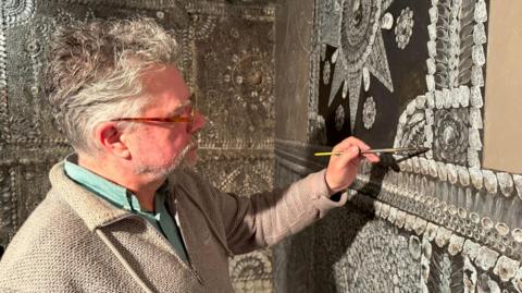 A man in a beige jumper and green shirt, who is wearing glasses, is restoring the Shell Grotto in Margate.