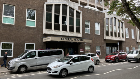 Cars drive past a brick building with the words General Hospital above an entrance. Passers by are walking in the street.
