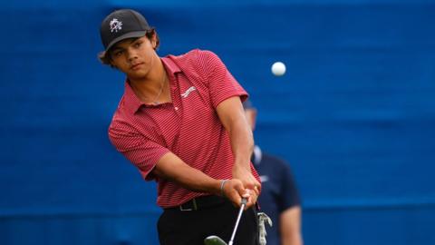 Charlie Woods is wearing a red and white striped shirt and a black hat. He is holding a golf club.