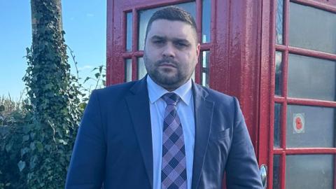 Alan Lewis in front of a red phone box. He has dark hair and beard, wearing a dark blue blazer, light shirt and check tie. A telephone pole covered in ivy can be seen to his right.