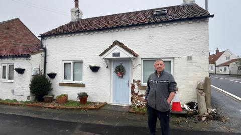 John Musgrave standing outside his home that has had its walls severely damaged after being hit by two cars