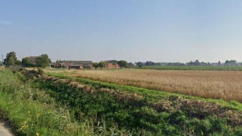 A view of the plot from the road.  There is a field with a crop growing in it at the moment, with some barns in the background.  The weather is sunny.