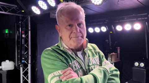 Steve Jason is standing on a small stage which has stage lighting on its ceiling and walls. Jason has short white hair and he is wearing a white T-shirt, and a green jacket which has white writing and stripes.