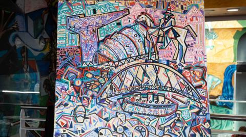 An abstract, expressionist painting of bright colours that show Glasgow's landmarks. There is a bridge, a crane and a statue painted amongst other abstract pictures. The painting is on an easel central to the photograph.