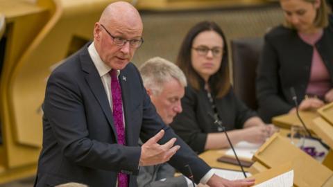 John Swinney, a bald man with glasses, speaks at a podium in the Scottish Parliament. He is wearing a dark suit, white shirt and purple tie, with his right hand gesticulating in front of his body. There are MSPs sat on benches behind him. 