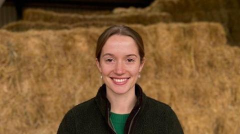 A smiling woman looking straight at the camera. She is standing in front of bales of hay and is wearing a green top and a black jumper.