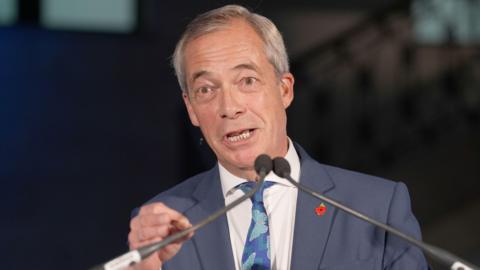 Nigel Farage giving a speech at Banking Hall in the City of London. He is wearing a blue suit and tie with a red poppy pin badge and speaking into a microphone.