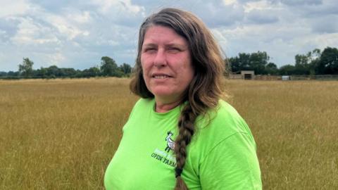 A woman stands alone in a wide field of hay beneath a cloud-filled sky. She wears a bright green T-shirt and has long, braided hair. Trees line the distant horizon.