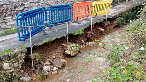 A section of collapsed footpath above a narrow river. Part of the wall supporting the tarmac footpath has been washed away and the sagging path is blocked off by plastic barrier fencing.