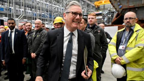Keir Starmer, wearing a dark suit, smiles as he looks off camera in BAE Systems facility in Govan, with men wearing high-vis and hard hats behind him 
