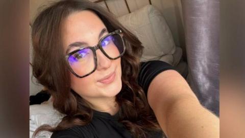 Alanis Sibley-Ware taking a selfie, wearing glasses and with brown curled hair. She is holding the camera up with her left arm and appears to have a bed with pillows and a purple curtain behind her. 