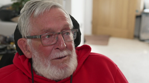 The image shows a close-up of an older person seated indoors. The person has short grey hair and a full grey beard and is wearing rectangular glasses and a red hooded sweatshirt. The person’s head is turned slightly to the left, looking off-camera.