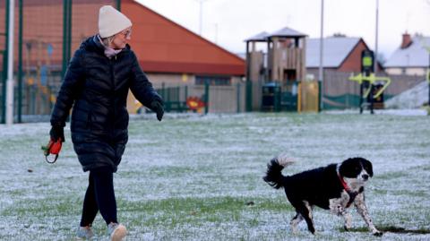 A woman wearing a hat and puffer jacket walks next to a dog. There's a dusting of snow on the ground