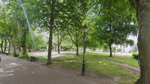 A green space in De Monfort Square with a number of mature trees and an ornate streetlight next to a pedestrian walkway
