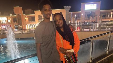 Osman, wearing a light grey t-shirt, stands with his arm round his mother, wearing an orange top and a black headscarf. It's night-time and there's a fountain behind them.