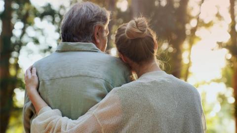 Two people from the back. One person has their arm around the other and is also resting their head on the other person’s shoulder. 