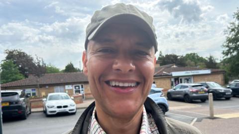 Clive Lewis is standing in front of a car park. He is wearing a green hat, white patterned shirt and green jacket. Behind him are a row of cars which are parked in front of a one-storey building.