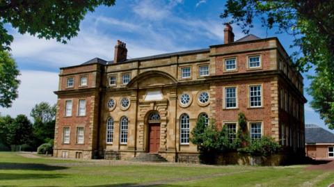Kirkleatham Museum is a grand three-storey brick and stone building in a grassy field. There is an arched window above the tall brown door. The other windows are a mix of circular and square designs. A lush green vine grows on the front wall.